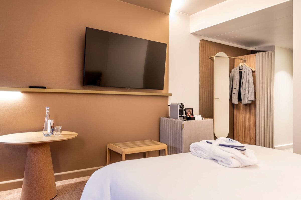 Hotelkamer met een bed, een aan de muur bevestigde televisie, een klein rond tafeltje met een glas en een bankje.
