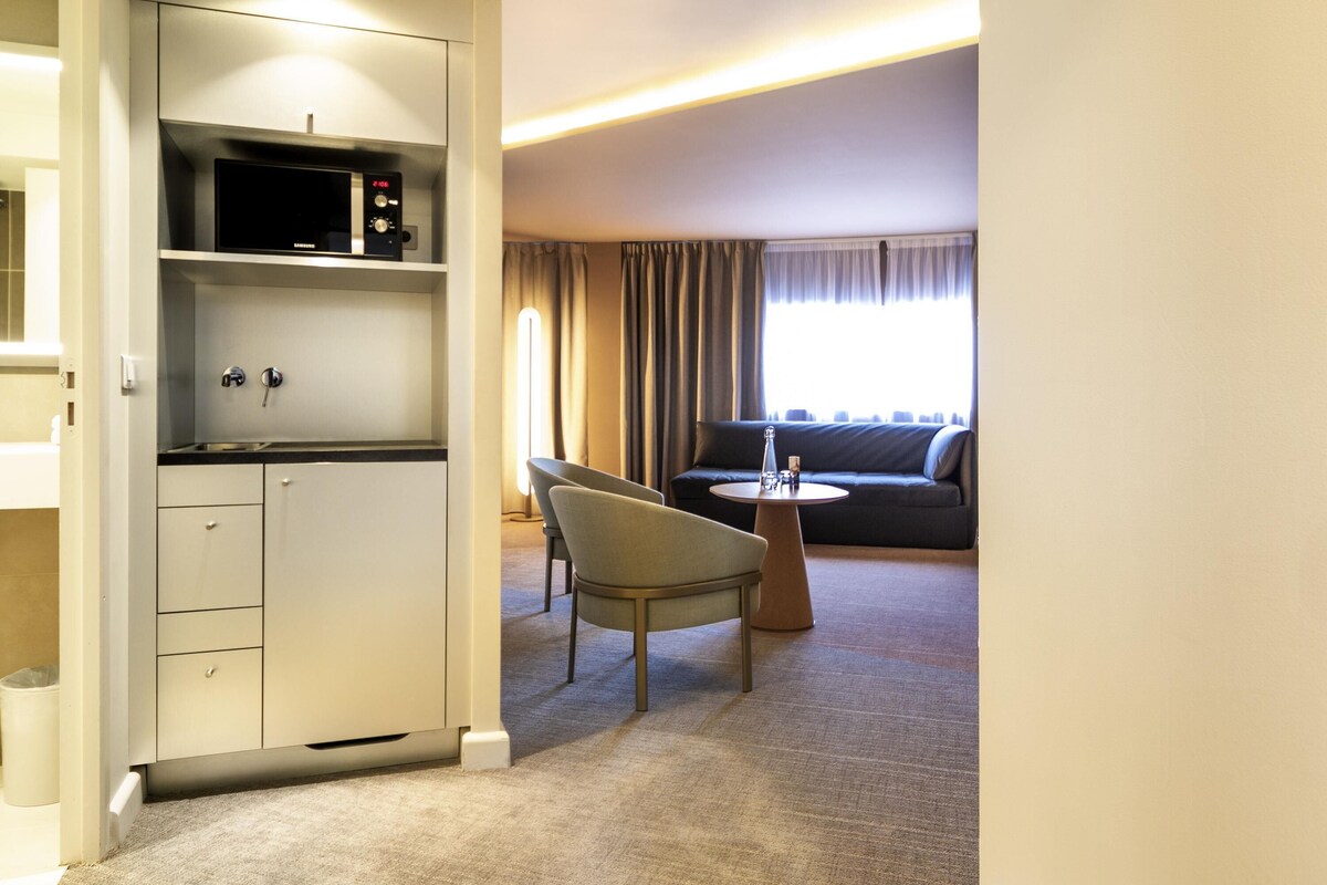 A modern hotel room with a kitchenette, a sofa, and a dining area.