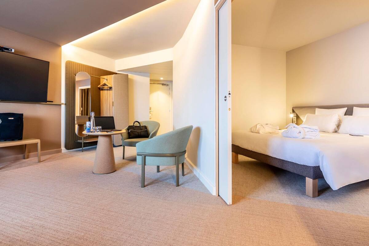 A modern hotel room with a bed, a desk with a laptop, a chair, and a TV.
