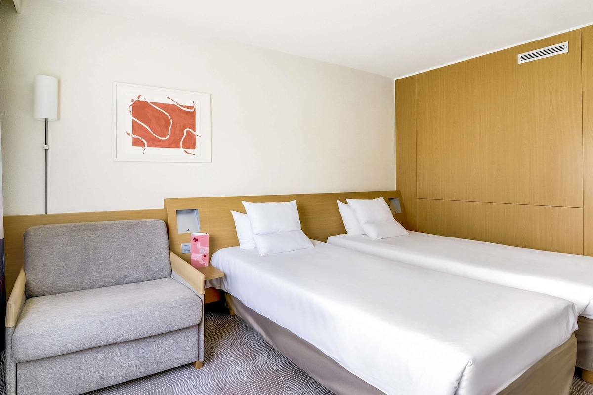 A hotel room with two single beds, a grey armchair, a bedside table with a lamp, and a framed picture on the wall.
