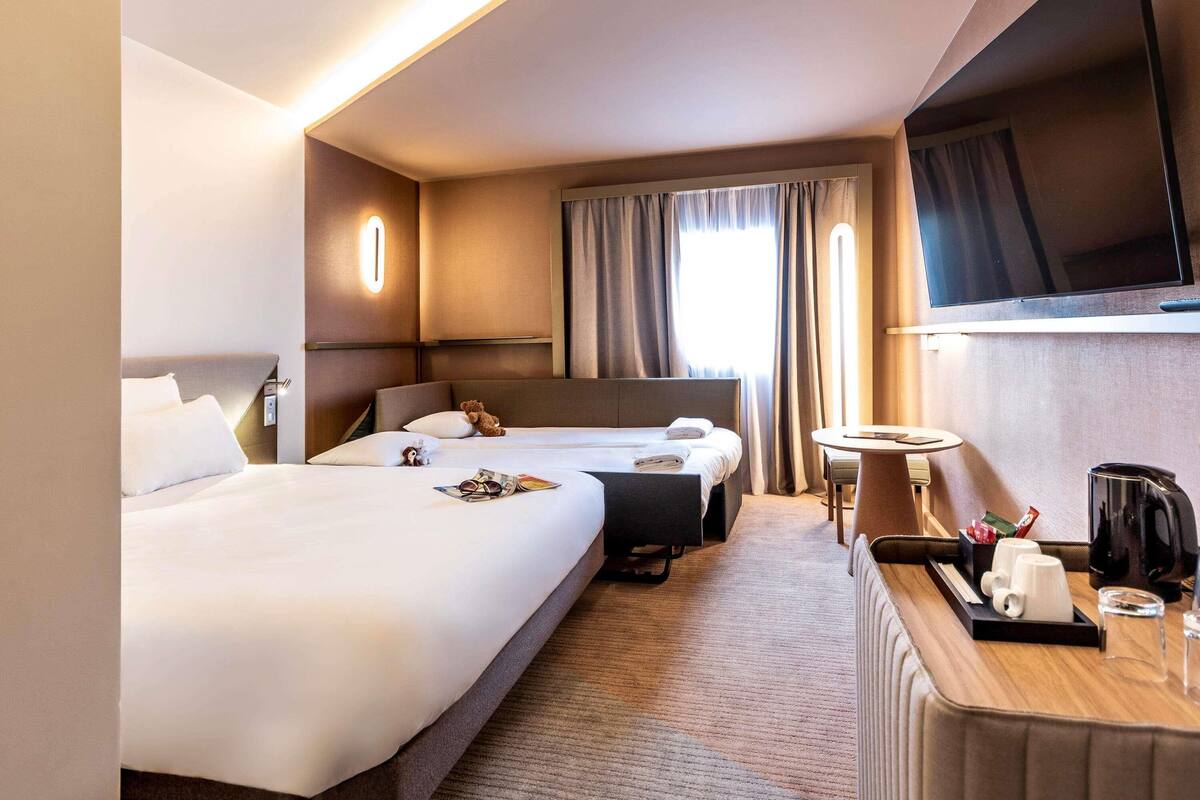 Een hotelkamer met twee bedden, een televisie, een bureau met koffiezetapparaat en een raam met gordijnen.