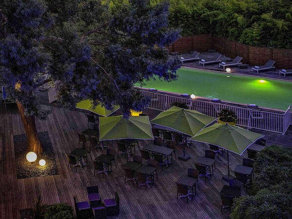 A deck with tables and chairs, a pool, and umbrellas. - Novotel Paris Orly Rungis (Rungis)