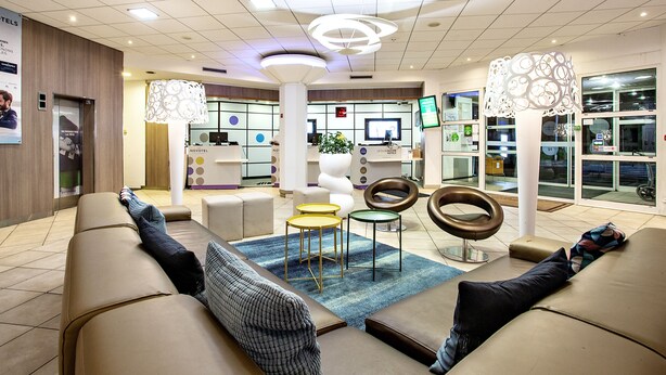 A modern hotel lobby with leather sofas, a reception desk, and a large window offering a view of the street.