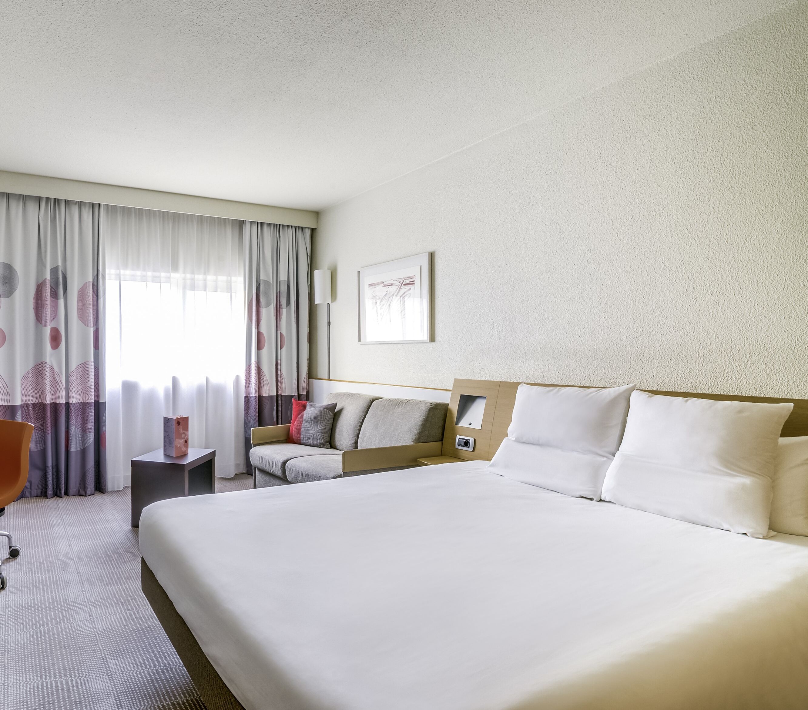 A hotel room with a large bed, a chair, a bedside table, and a window with curtains.