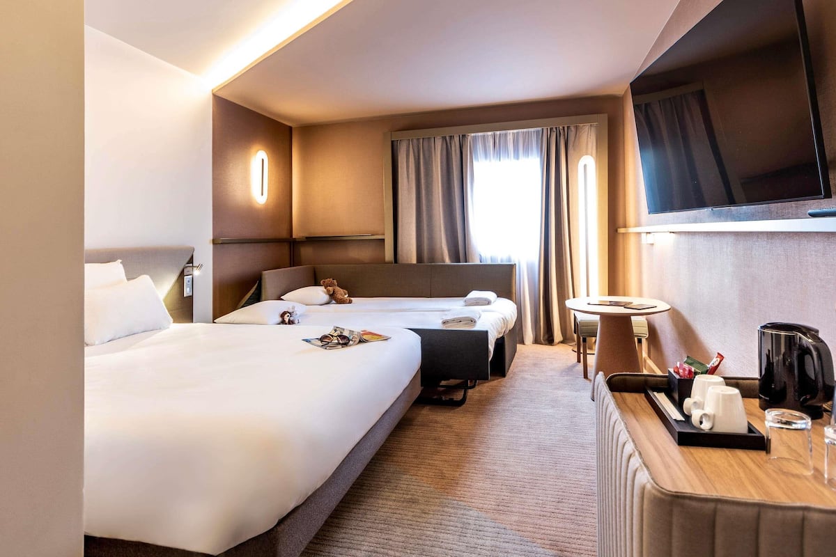 A hotel room with two beds, a TV, a desk with a coffee maker, and a window with curtains.