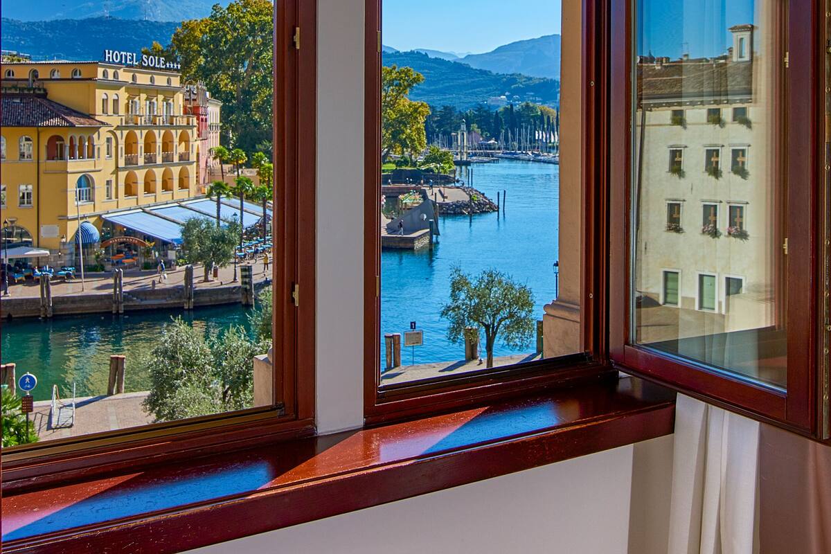 Ein Hotelzimmer mit einem großen Bogenfenster mit Blick auf ein Gewässer, einen Steg und ein Gebirge.