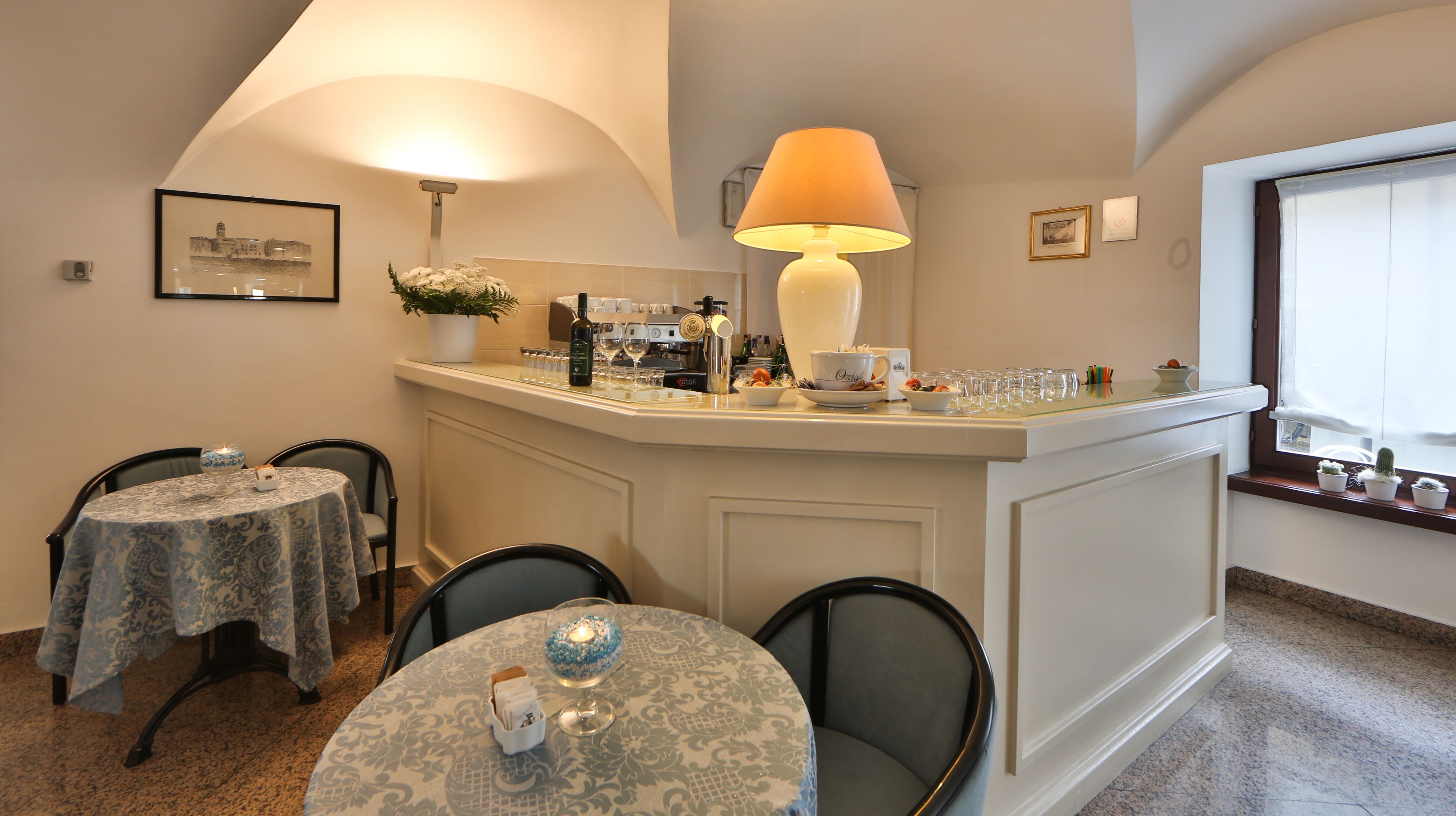 A dining area with a bar, two round tables with patterned tablecloths, chairs, a lamp, and a potted plant.