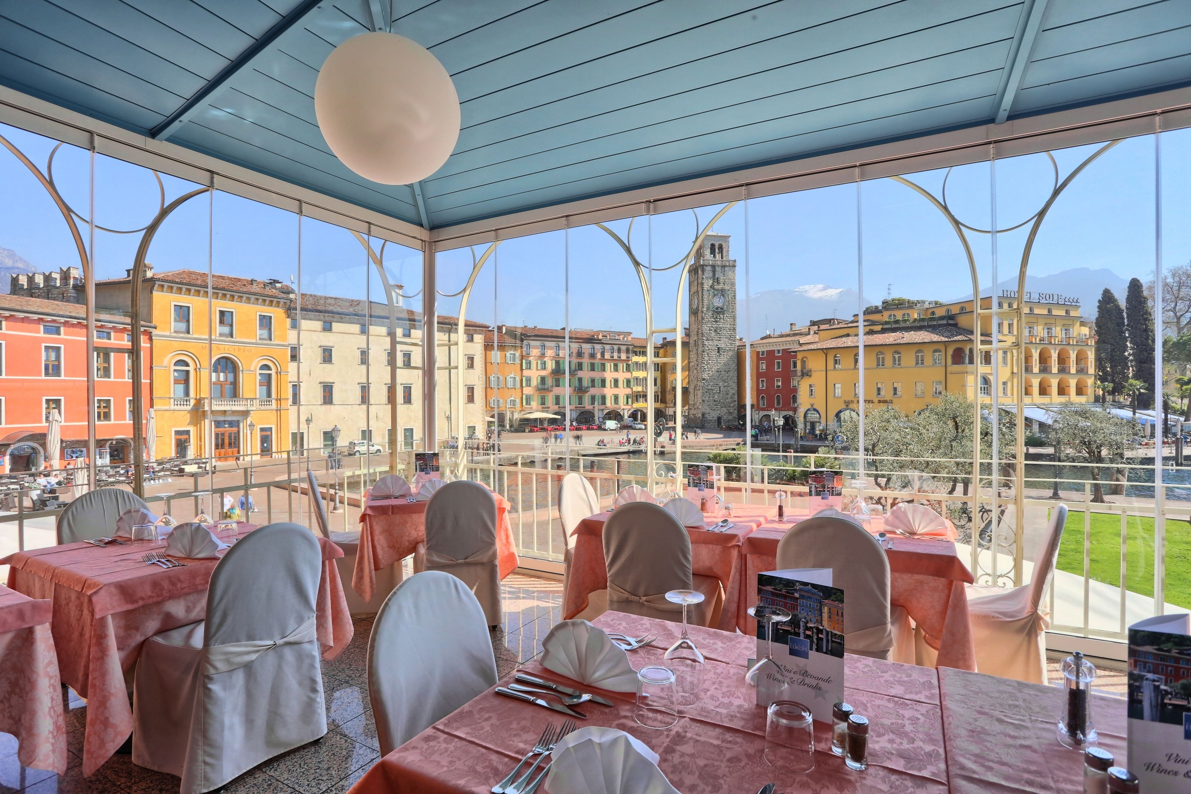 A restaurant with tables set for dining, large windows offering a view of the cityscape, and a clear sky.