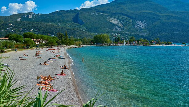 Ein Strand mit klarem türkisblauem Wasser, einer felsigen Küste und einem Berg im Hintergrund.