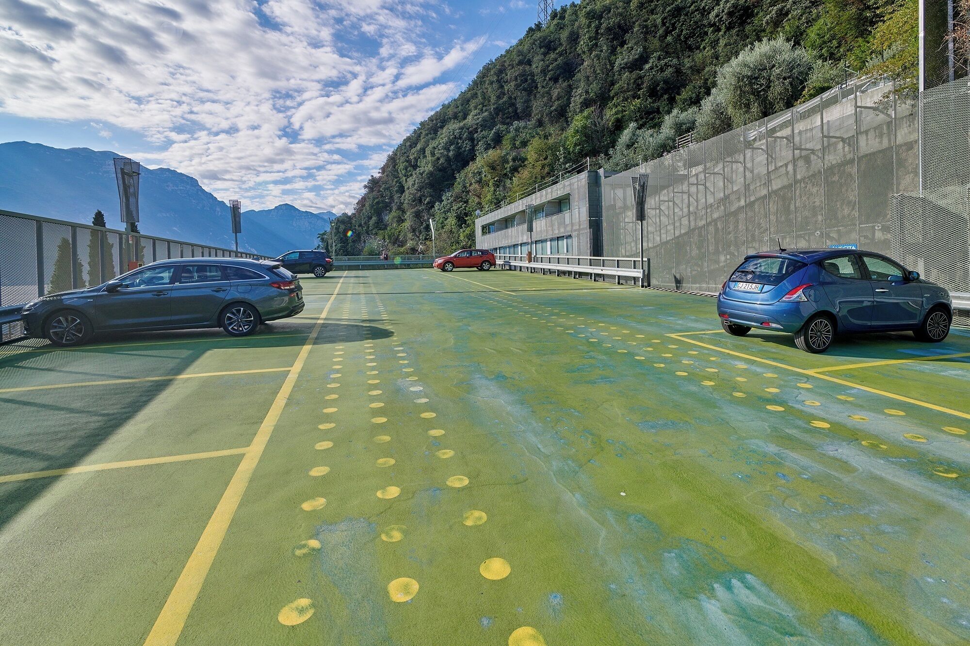 Un espace de stationnement avec des voitures, une surface verdoyante avec des marquages jaunes et un paysage montagneux en arrière-plan.