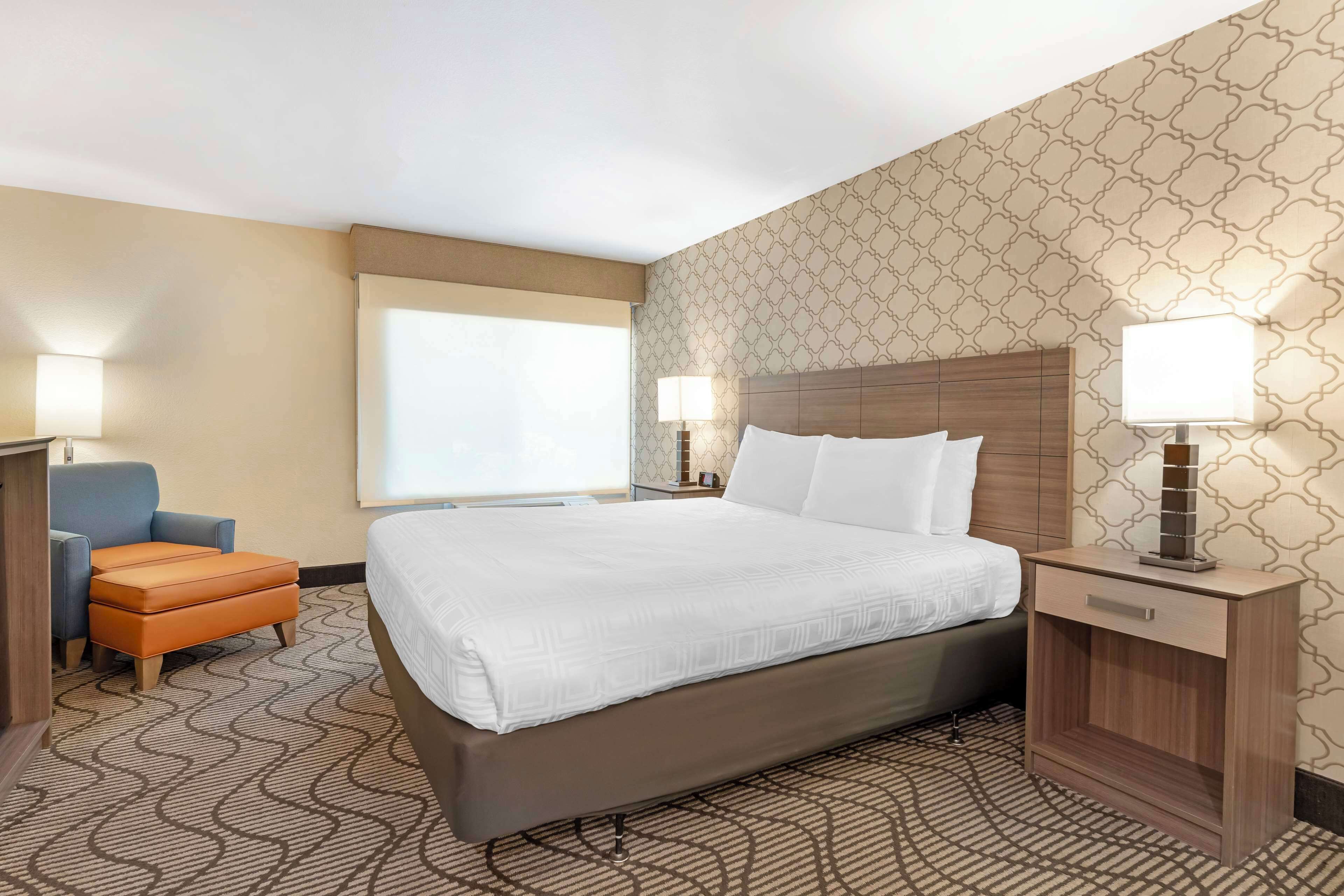 A hotel room with a large bed, a nightstand with a lamp, a blue armchair, and a patterned carpet.