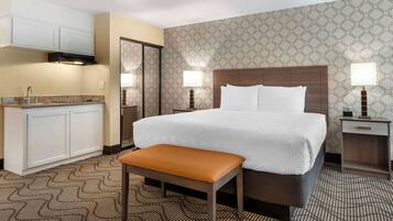 A hotel room with a bed, a bench, a nightstand, a lamp, a small table, a sink, and cabinets.