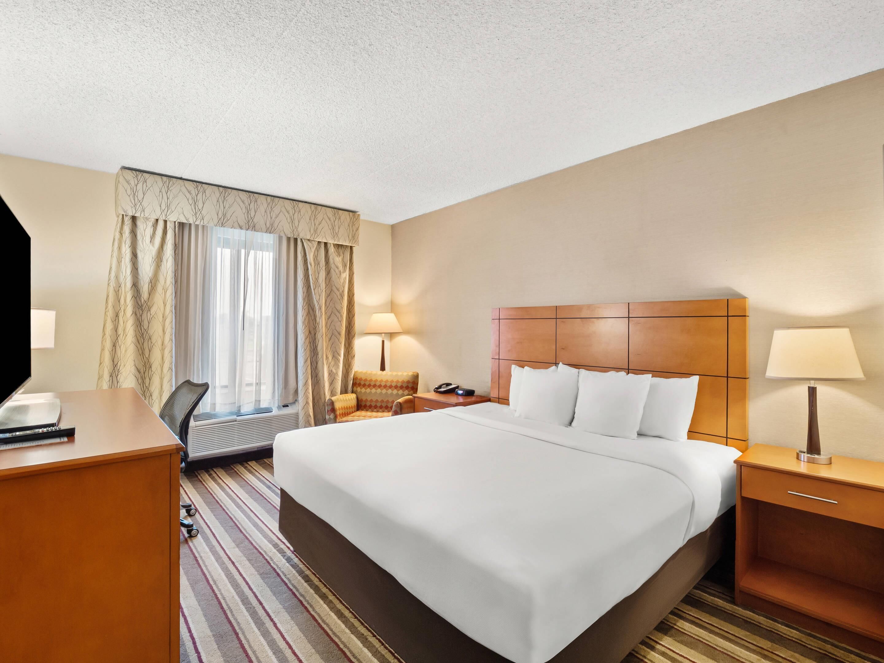 A hotel room with a large bed, a desk, a chair, a nightstand, and a window with curtains.