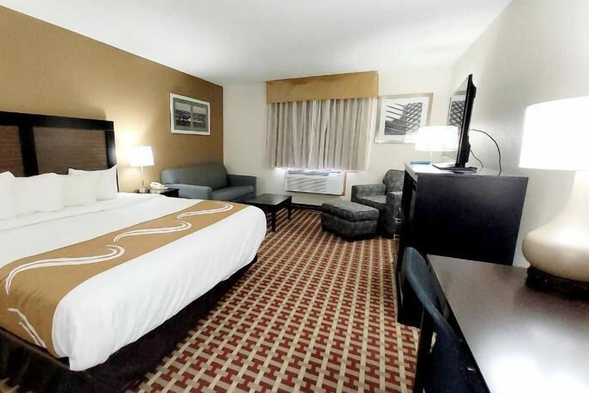 Quarto de hotel com uma cama grande, escrivaninha, cadeira, televisão e janela com cortinas.