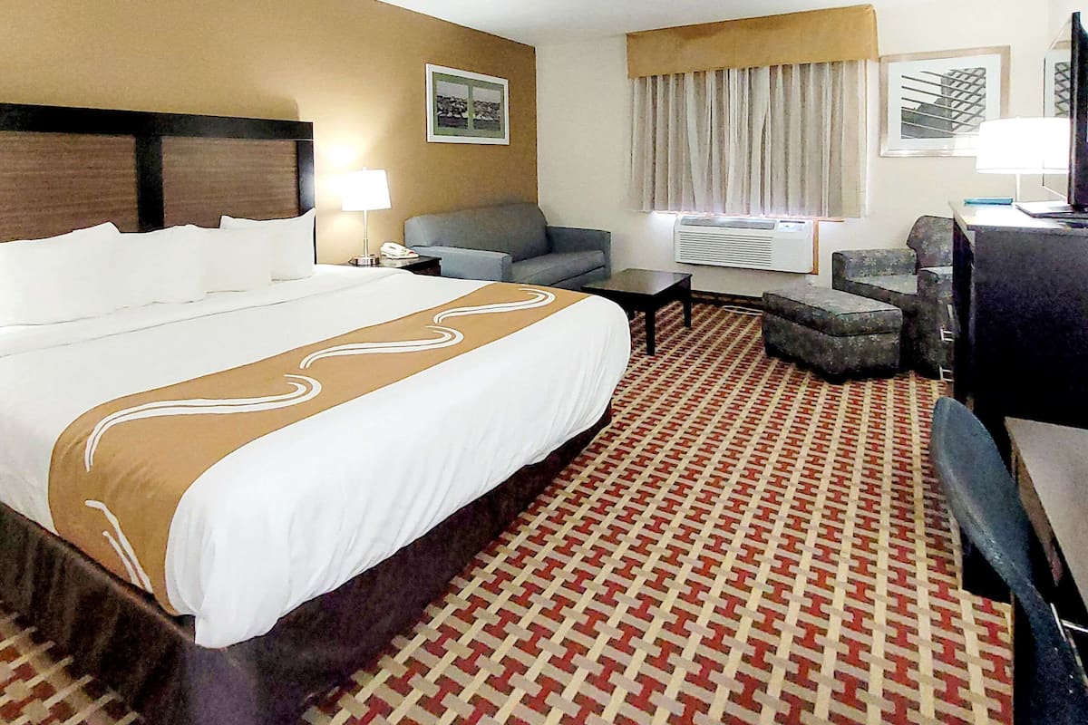 Quarto de hotel com uma cama grande, uma poltrona cinza, uma pequena escrivaninha e um tapete estampado.