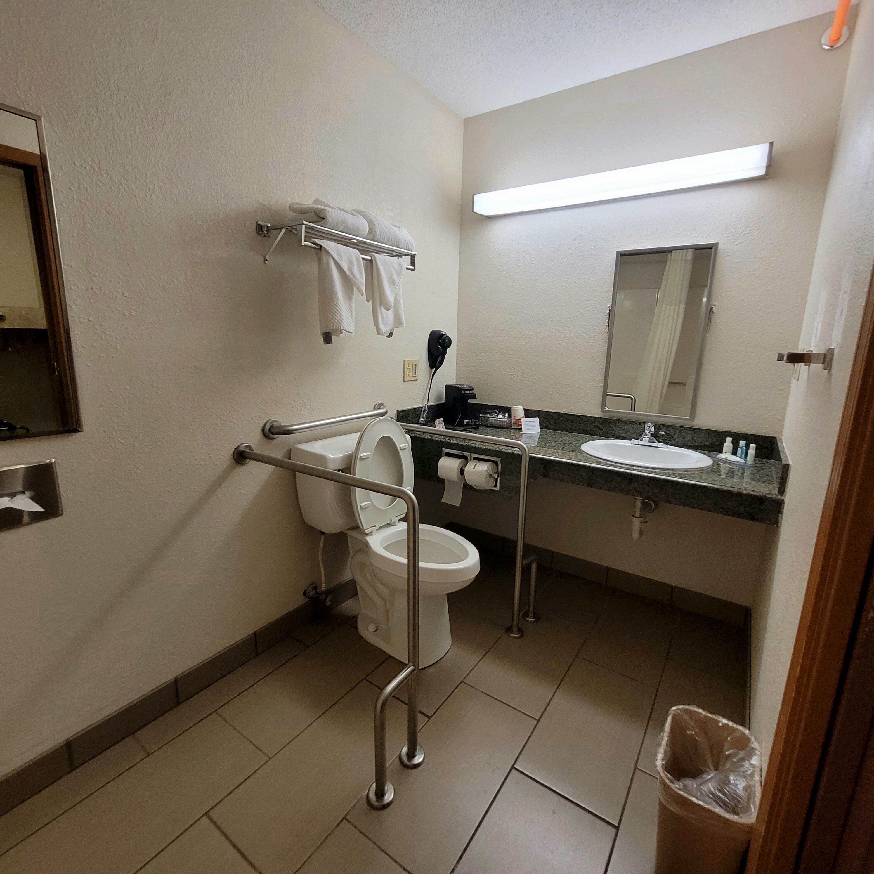 A standard hotel bathroom with a toilet, sink, and mirror. There is a towel rack above the toilet and a trash can in the corner.