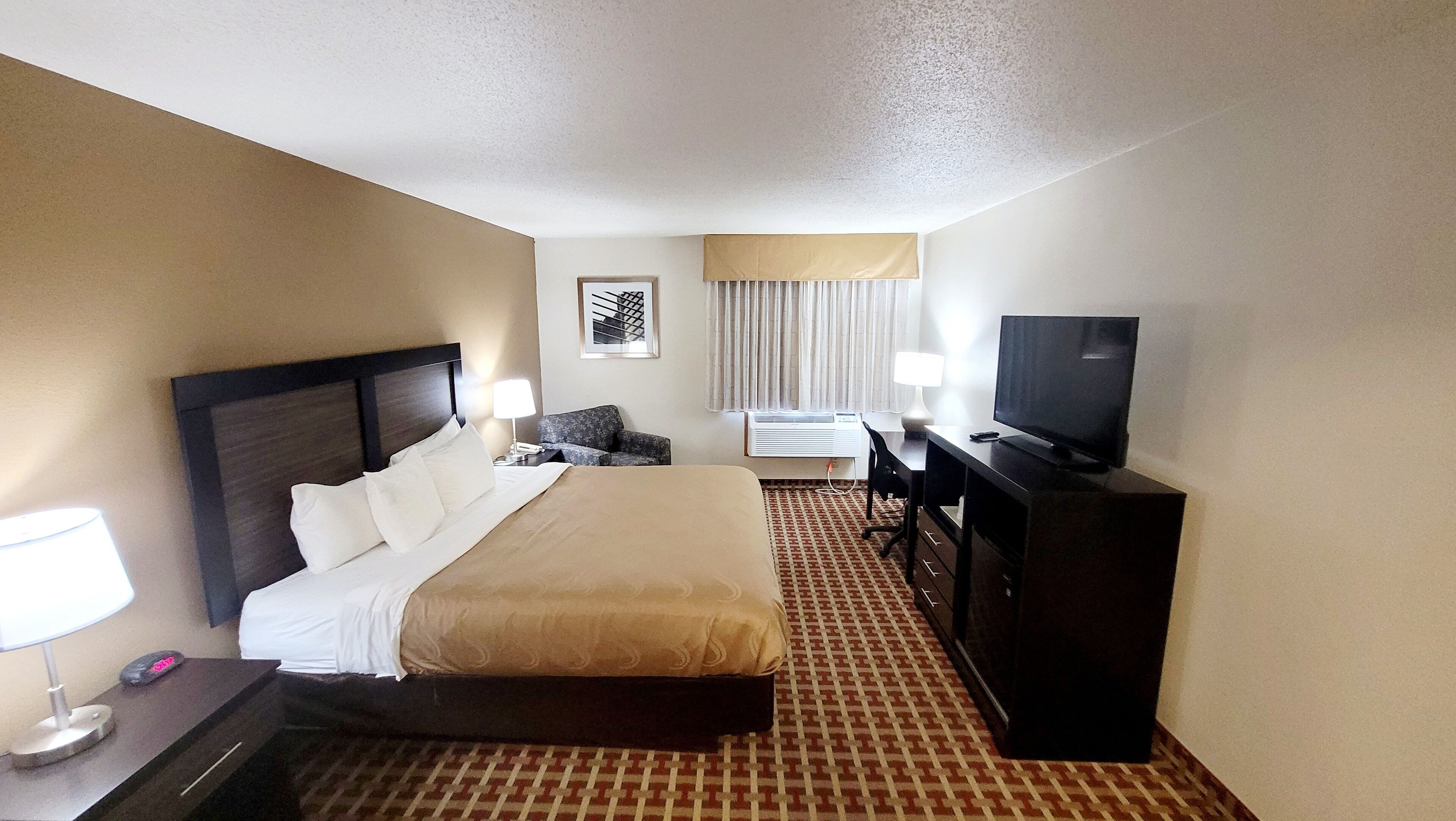 A hotel room with a bed, a desk, a television, and a patterned carpet.