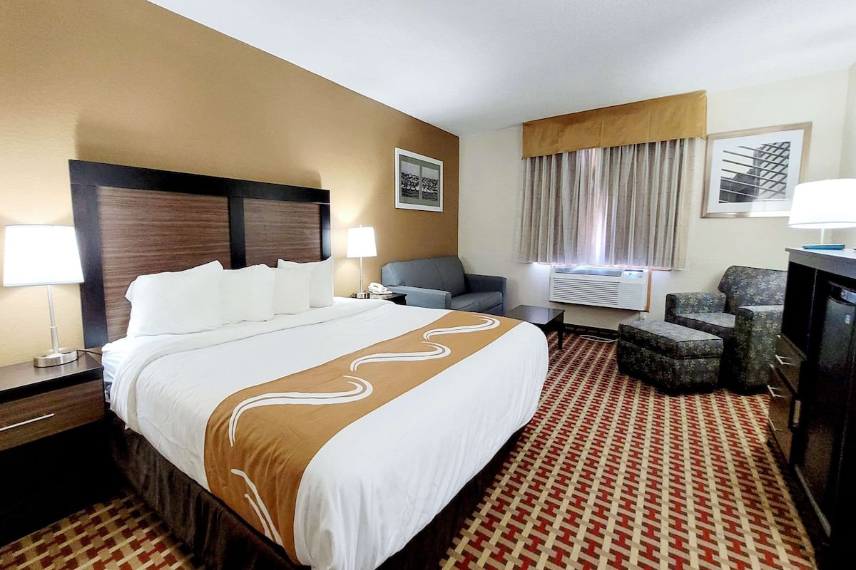 Quarto de hotel com uma cama grande, uma escrivaninha com abajur, uma cadeira, ar condicionado e televisão.