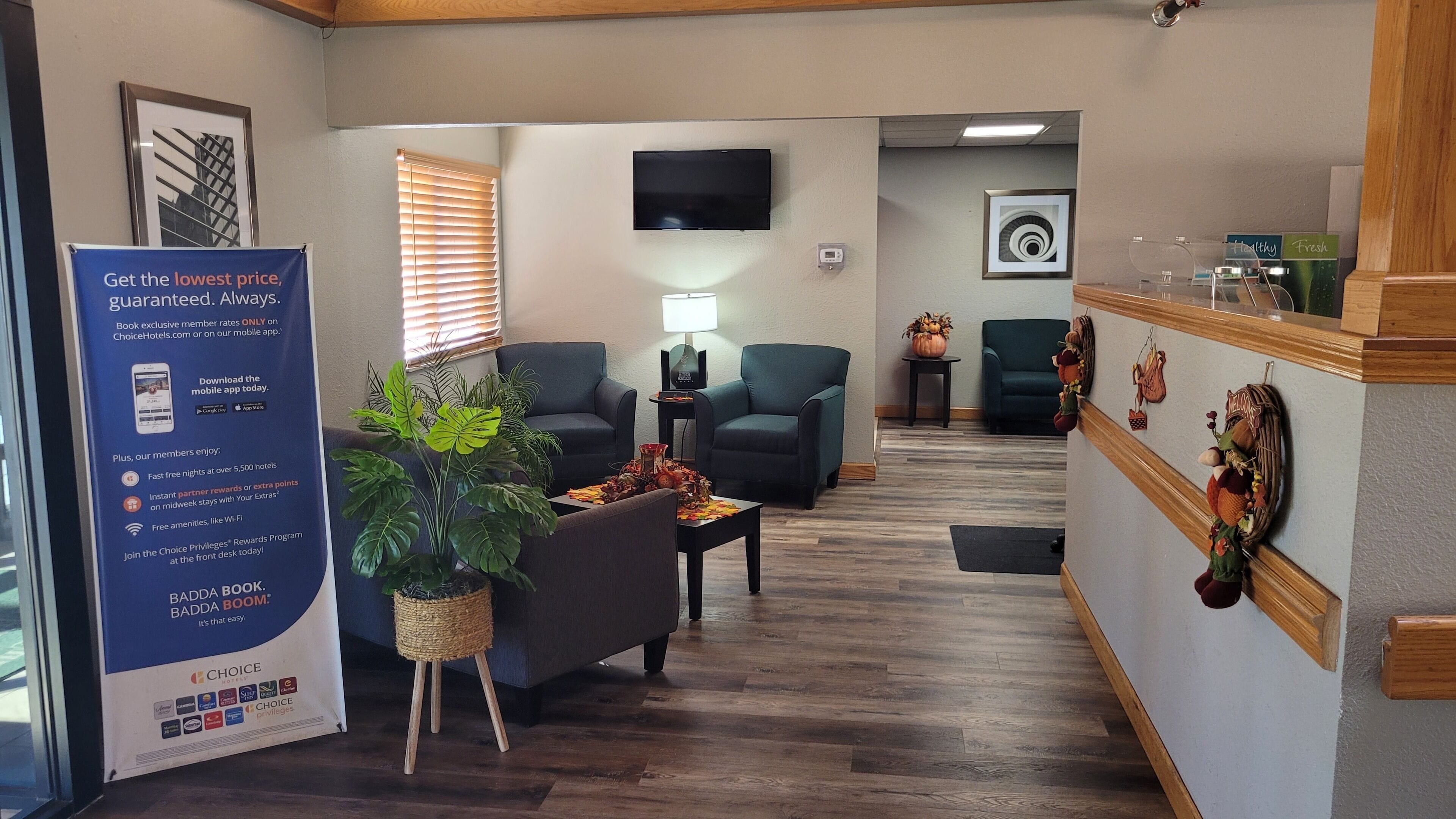 A hotel lobby with a reception desk, two armchairs, a coffee table, a potted plant, a TV, and a wall with a welcome sign.