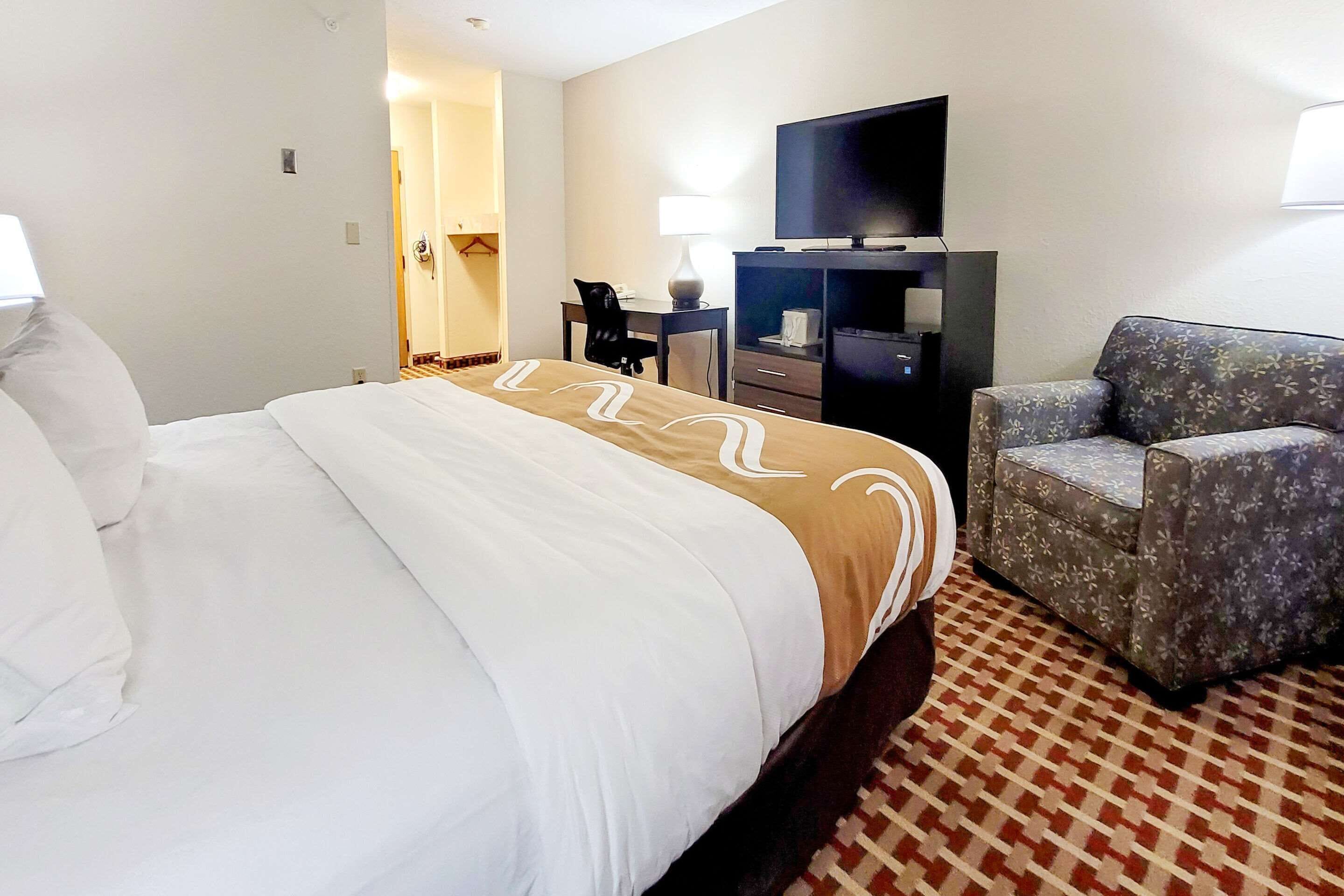 A hotel room with a large bed, a desk with a chair, a television, and a patterned armchair.