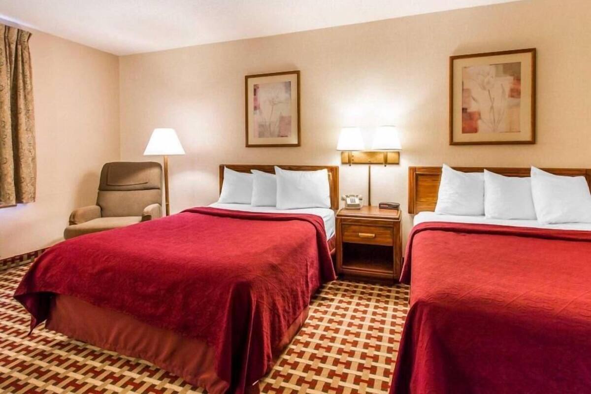 Quarto de hotel com duas camas, uma cadeira, um abajur e duas imagens emolduradas na parede.