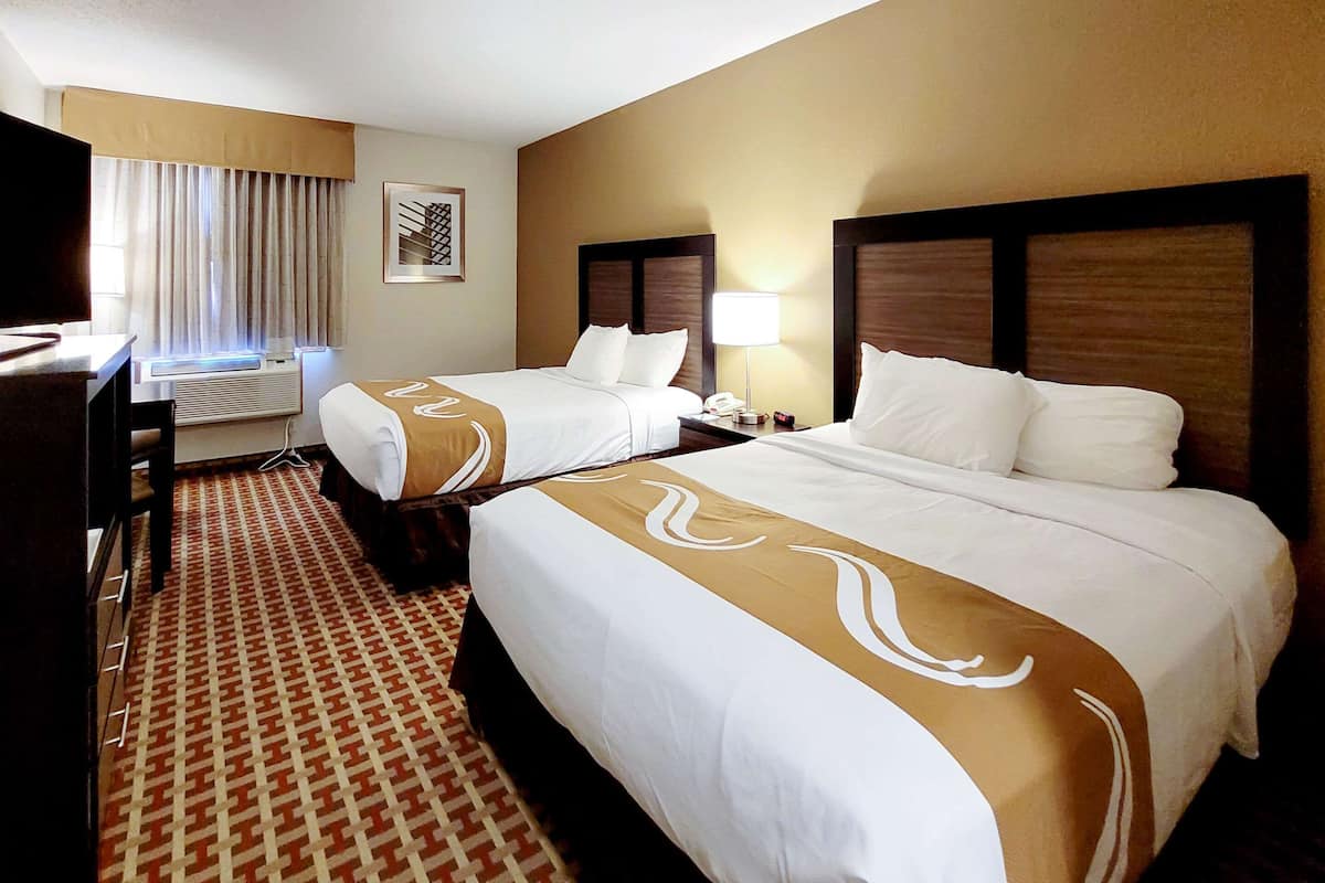 Quarto de hotel com duas camas, uma escrivaninha, uma cadeira, uma televisão e uma janela com cortinas.