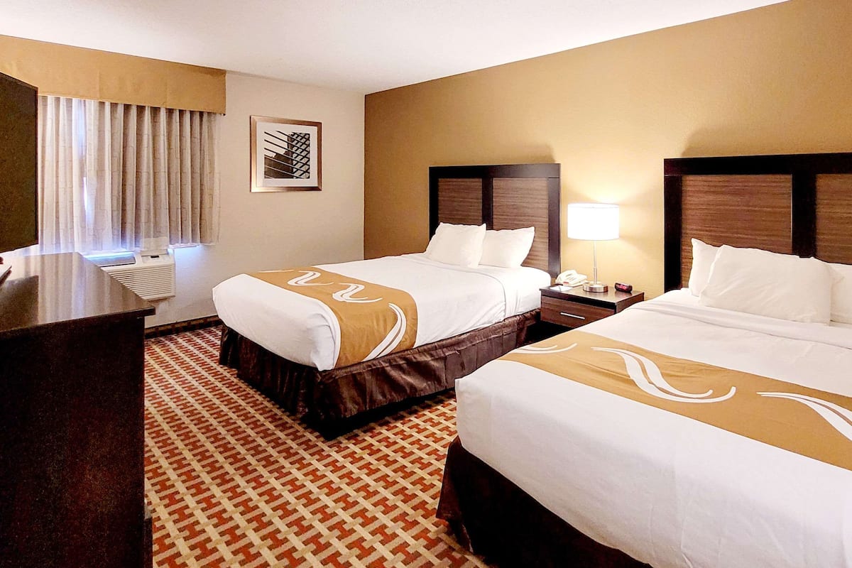 Quarto de hotel com duas camas, uma escrivaninha, um abajur e um tapete estampado.