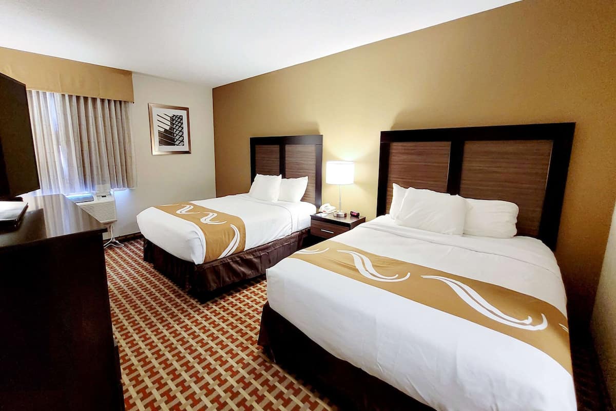 Quarto de hotel com duas camas, uma escrivaninha, uma luminária e um tapete estampado.