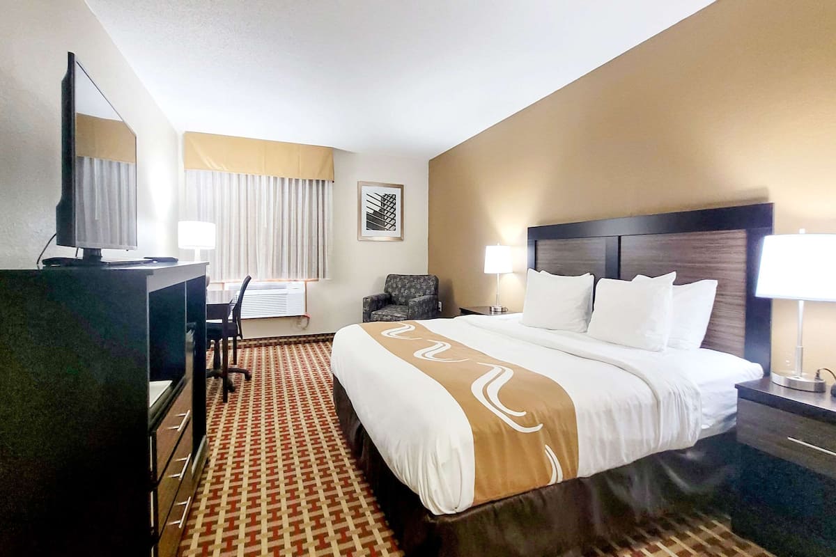 Quarto de hotel com uma cama grande, uma escrivaninha com computador, uma cadeira e uma televisão.