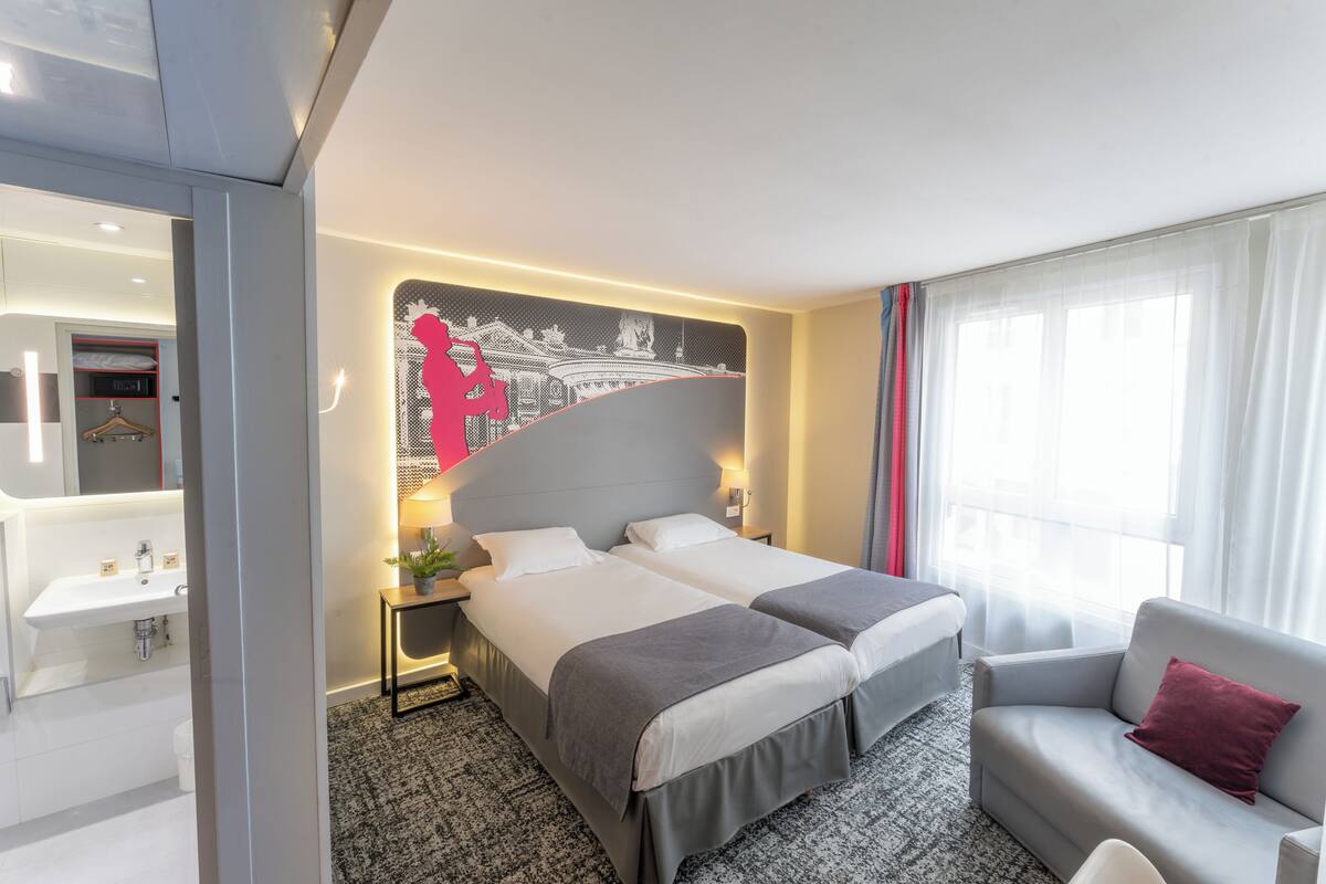 Une chambre d’hôtel avec deux lits, un canapé, un bureau avec une lampe, une grande fenêtre avec des rideaux et une salle de bain avec un lavabo et un miroir.