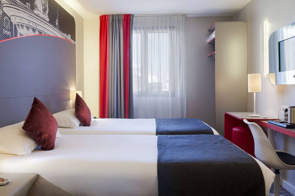 Une chambre d’hôtel avec deux lits, un bureau, une chaise, une télévision et une grande fenêtre avec des rideaux légers.