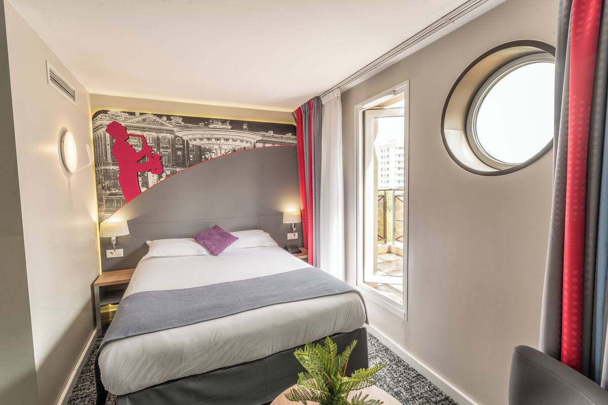 Une chambre d’hôtel comprenant un lit, une table de chevet, une lampe, une fenêtre avec des rideaux, une lucarne ronde et un mur orné d’un motif graphique.
