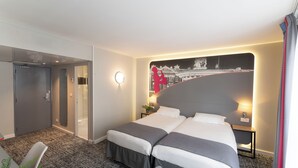 A hotel room with two beds, a large headboard with a cityscape mural, a small bedside table with a lamp, and a door leading to a bathroom. - Hotel Inn Design Paris Place d'Italie (Paris)