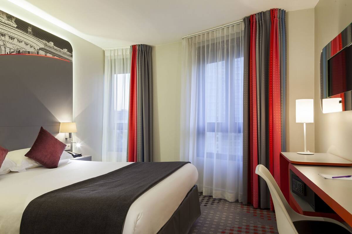 Une chambre d’hôtel avec un lit, un bureau, une chaise, une lampe et une grande fenêtre avec des rideaux.