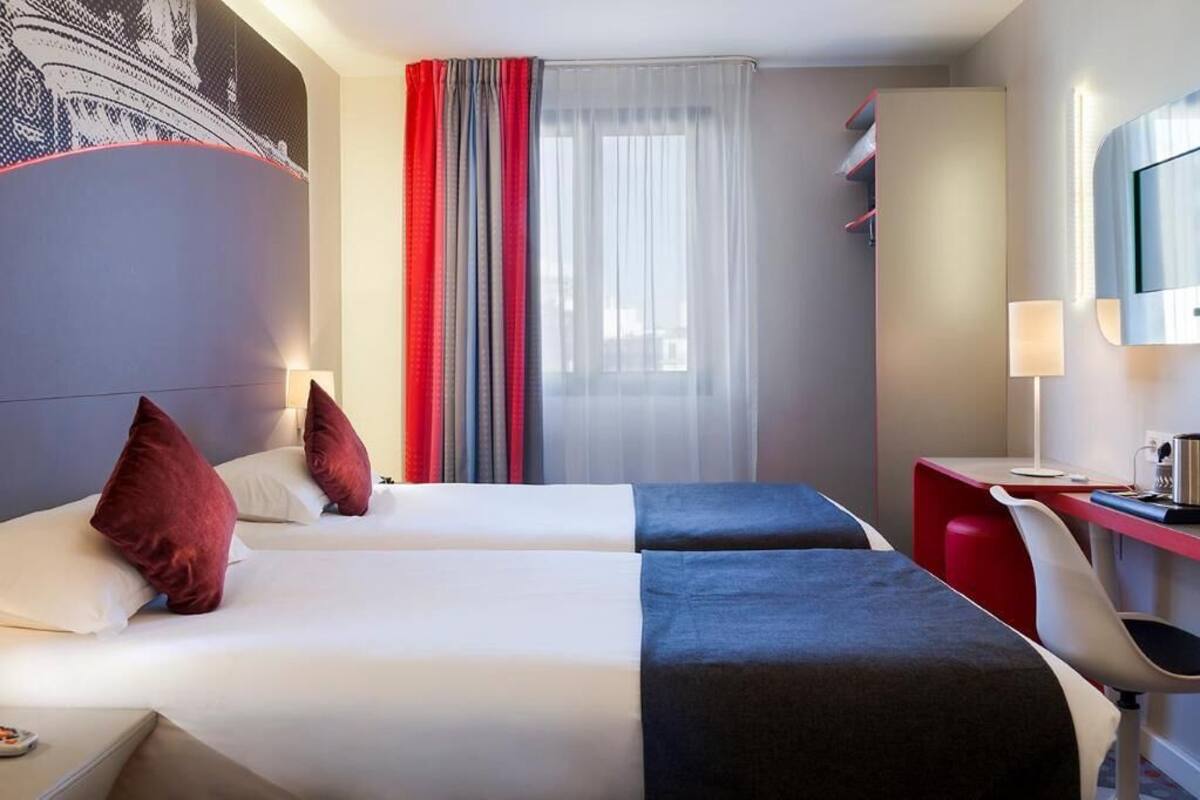 Une chambre d’hôtel avec deux lits, un bureau, une chaise, une télévision et une fenêtre avec des rideaux.