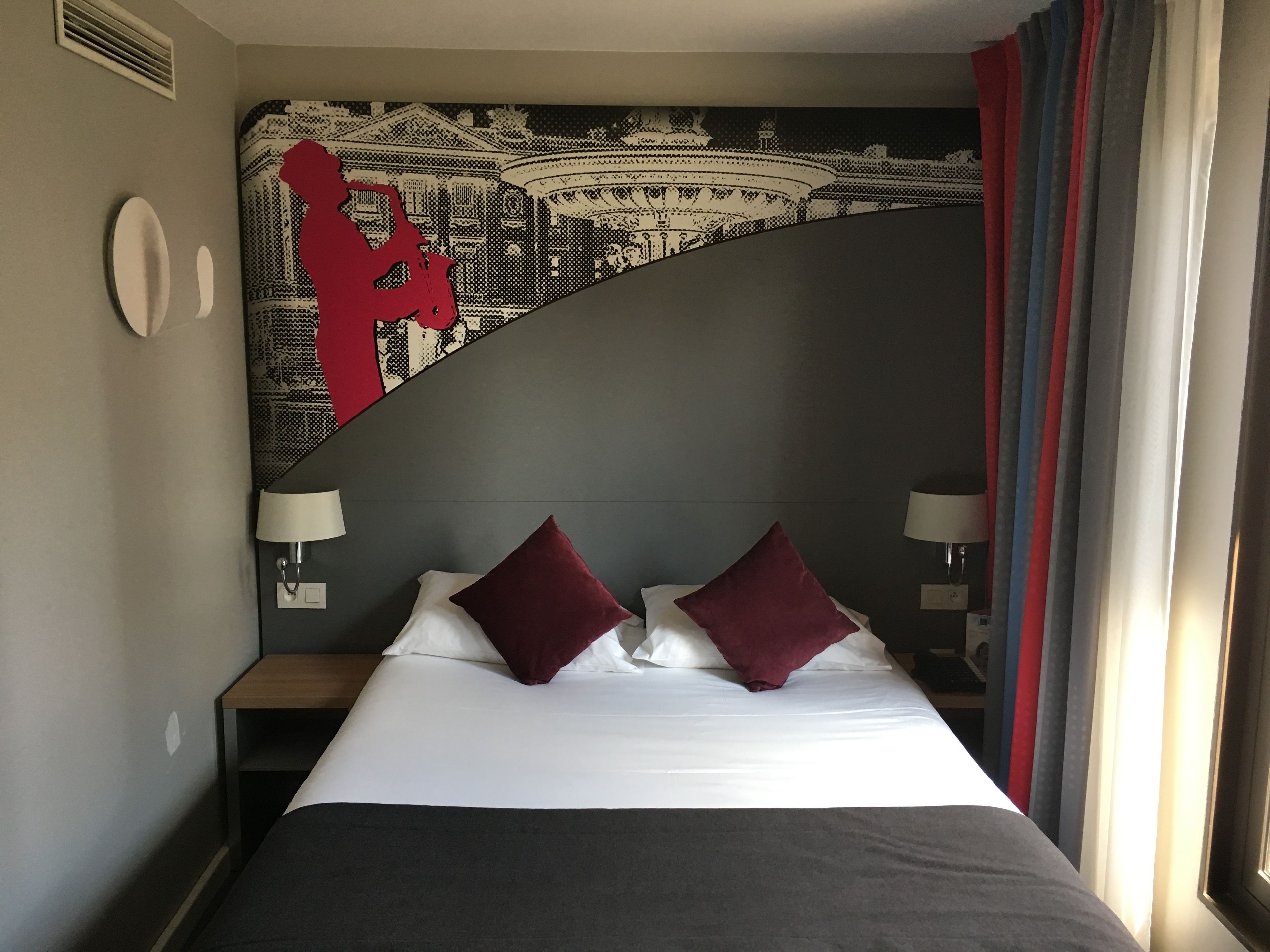 A hotel room with a bed, two pillows, a nightstand, a lamp, and a wall tapestry.