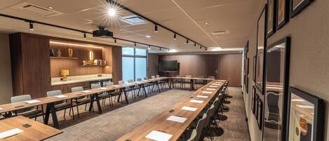 A conference room with long tables, chairs, a projector, and a large screen.