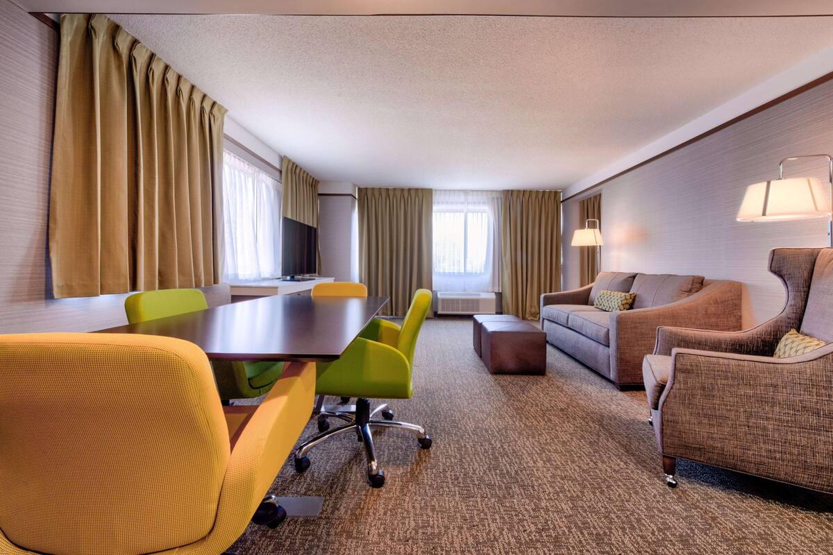 A hotel room with a conference table, yellow and green chairs, a sofa, and a TV.