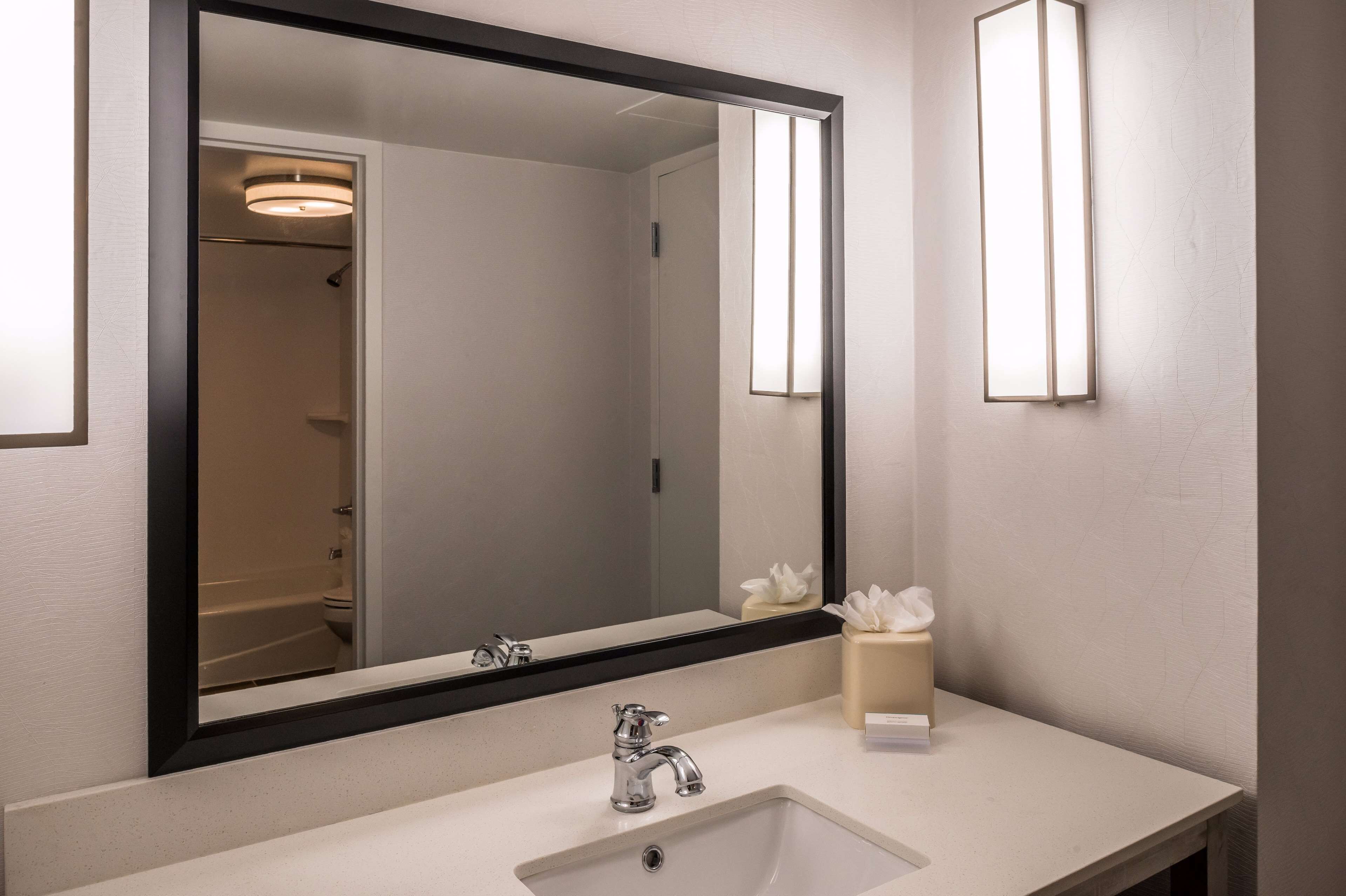 A bathroom with a large mirror, a sink, a towel dispenser, and a shower visible in the background.