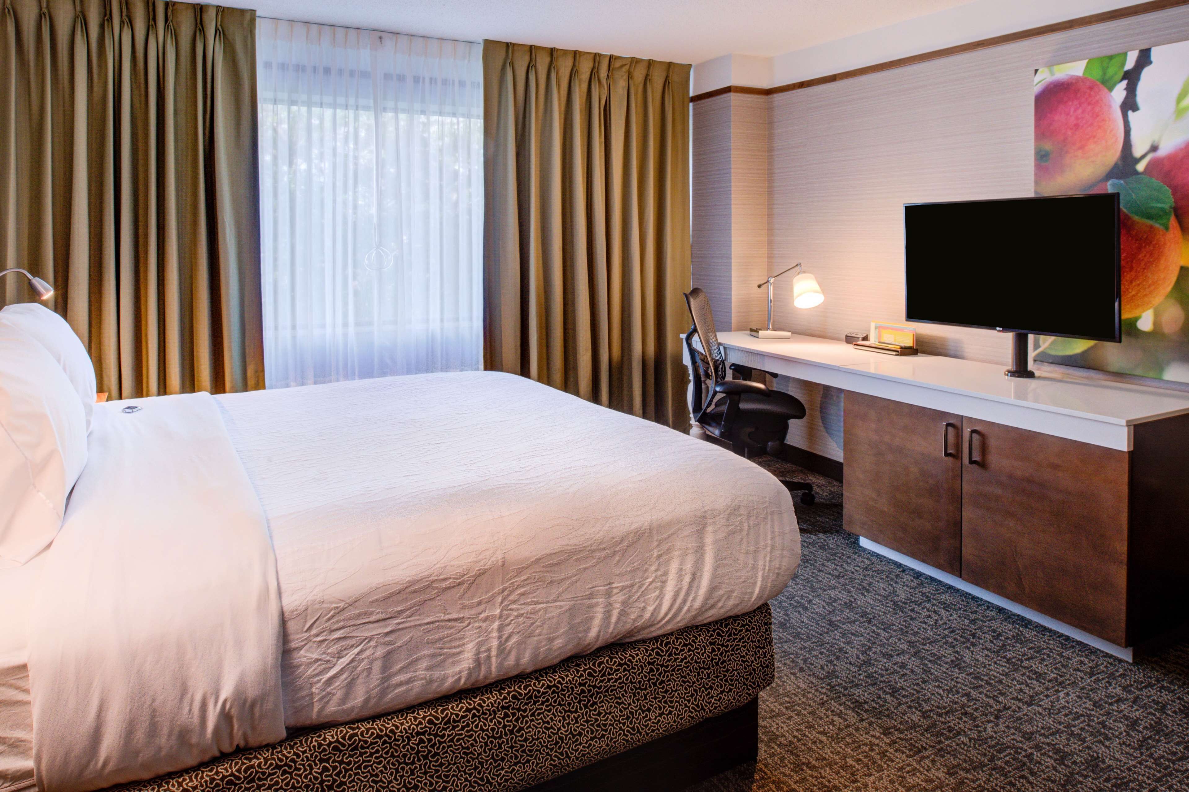Une chambre d’hôtel avec un grand lit, un bureau avec une chaise, une télévision et un tableau représentant des fruits accroché au mur.
