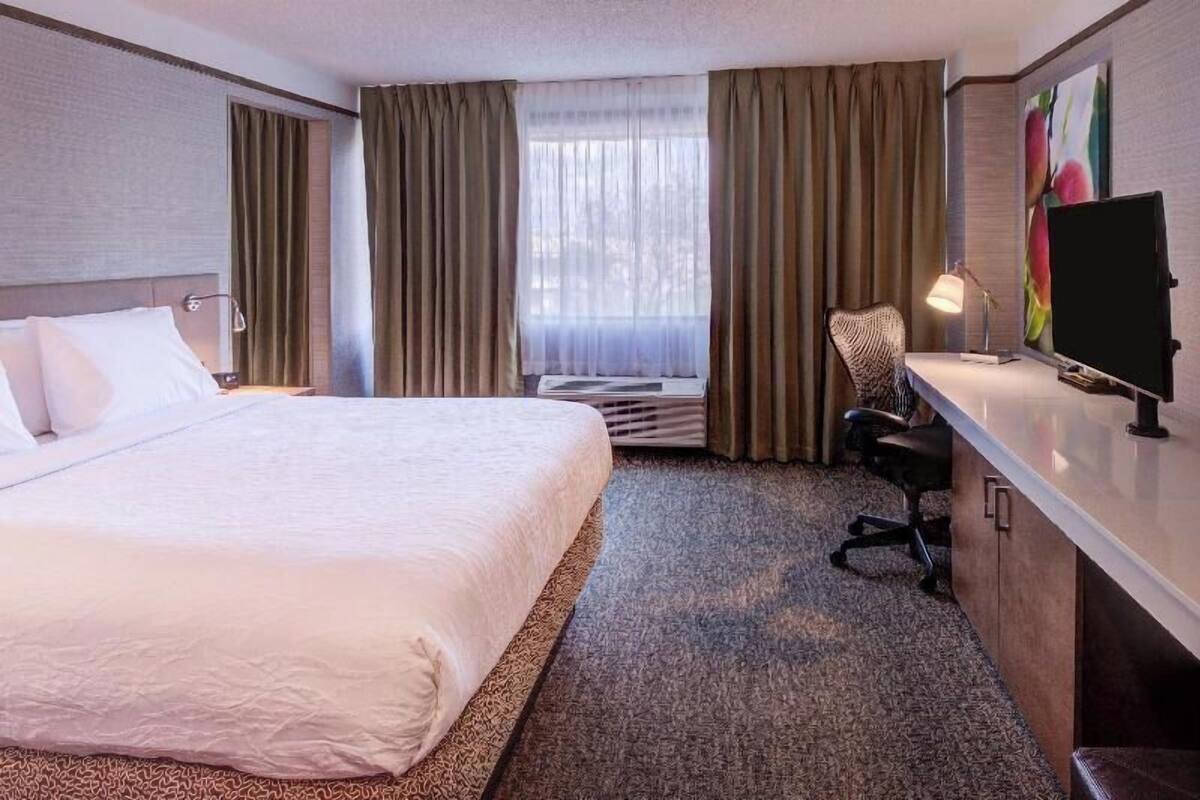 A hotel room with a large bed, a desk with a computer, a chair, and a window with curtains.