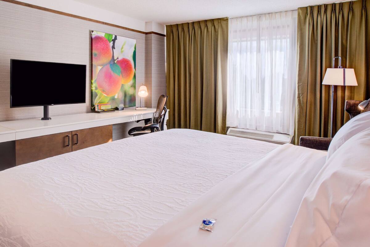 A hotel room with a large bed, a desk with a flat-screen TV, a painting, a lamp, and a window with curtains.
