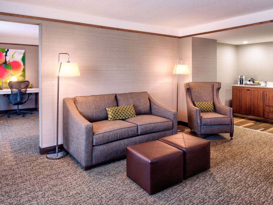 A hotel room with a sofa, armchair, ottoman, desk, chair, lamp, and a kitchenette area.