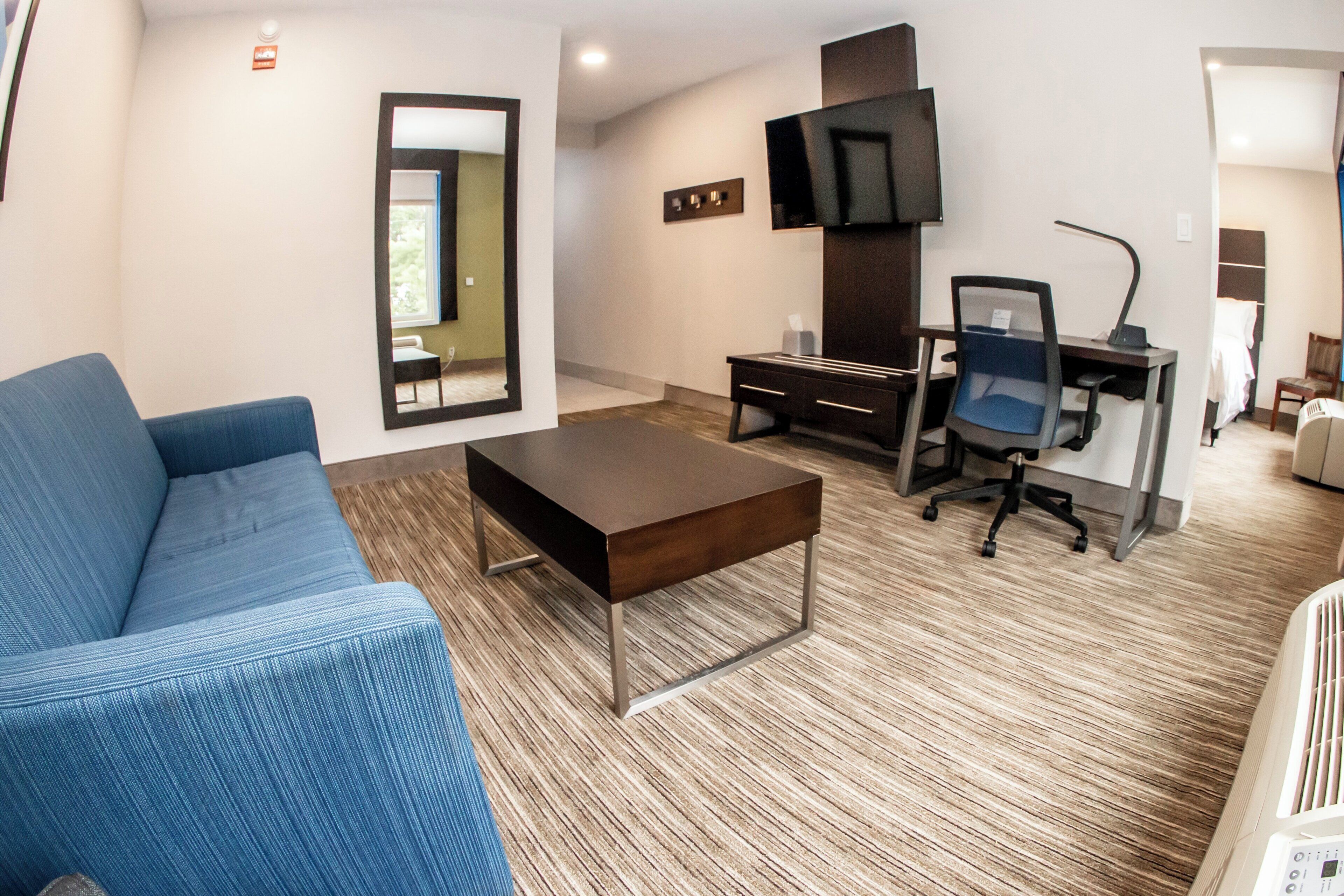 A hotel room with a blue sofa, a wooden coffee table, a desk with a chair, a flat-screen TV mounted on the wall, and a bed in the background.