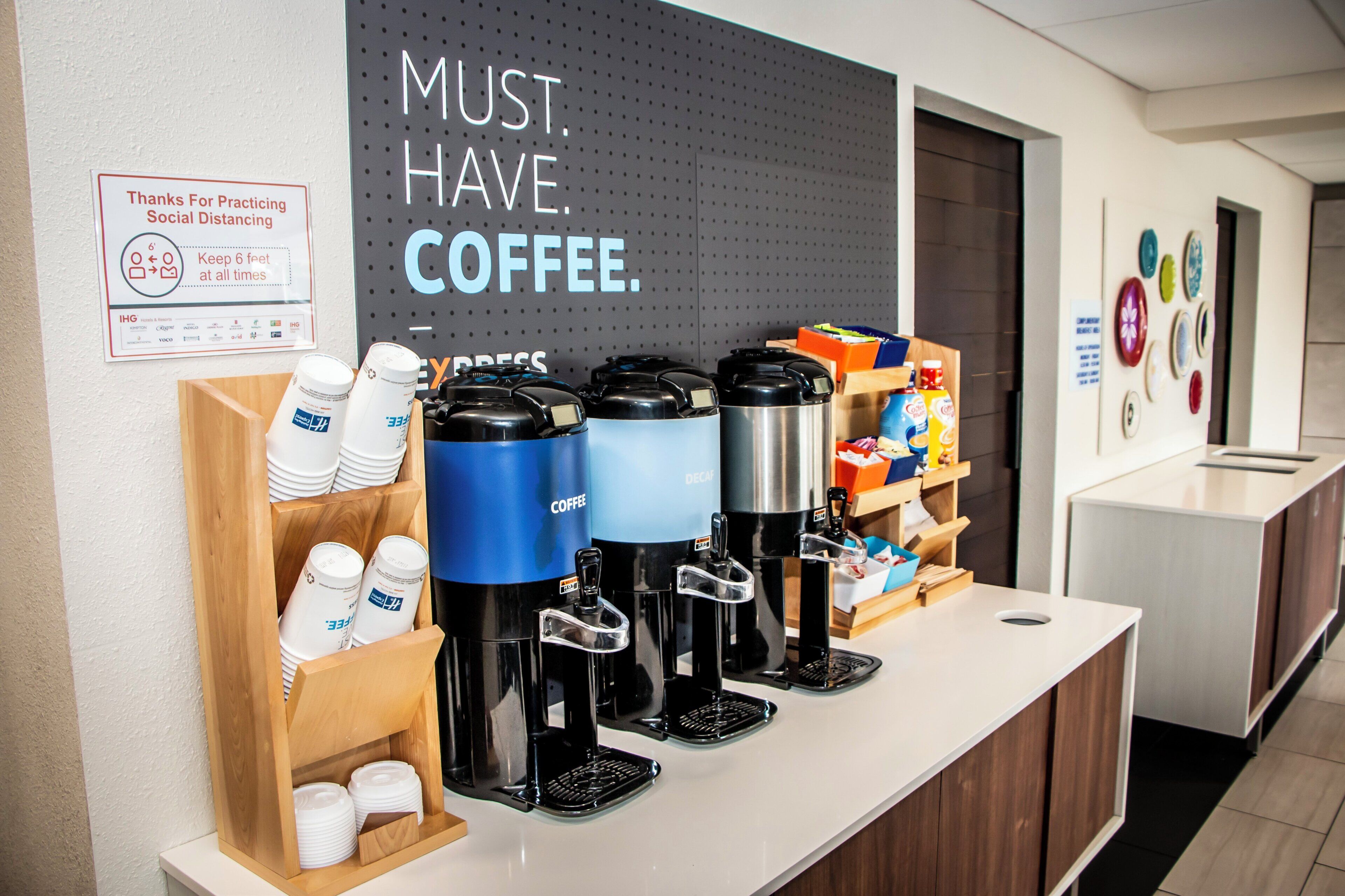 A coffee station with multiple coffee dispensers, a sign encouraging social distancing, and a wall with a motivational message.