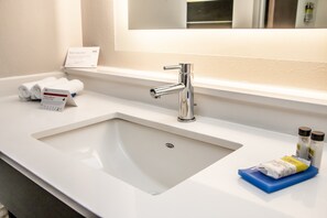 Un baño de hotel con un lavamanos blanco, un espejo encima y artículos de aseo en la encimera.