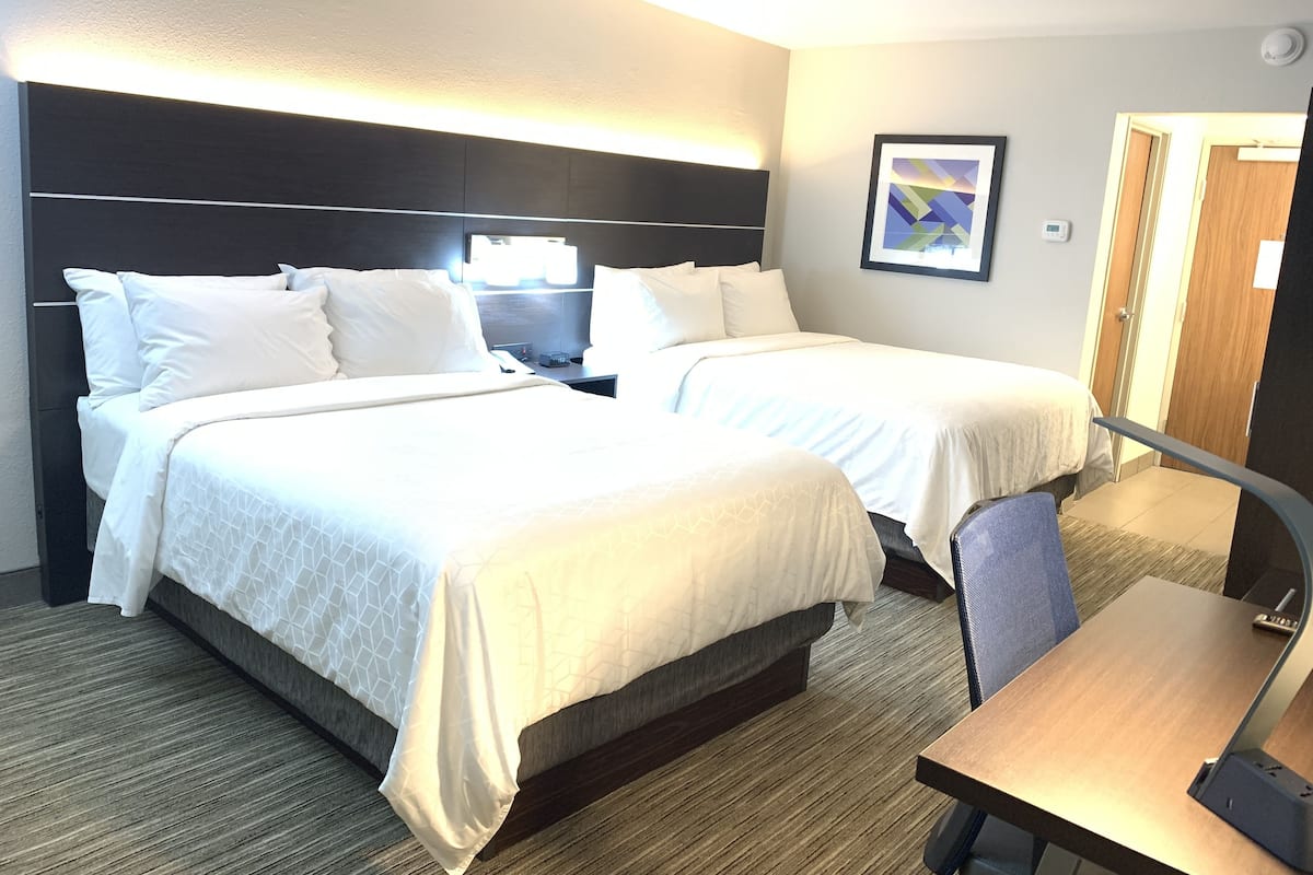 Habitación de hotel con dos camas, un escritorio, una silla, una mesita de noche, una lámpara, un cuadro enmarcado y una puerta.