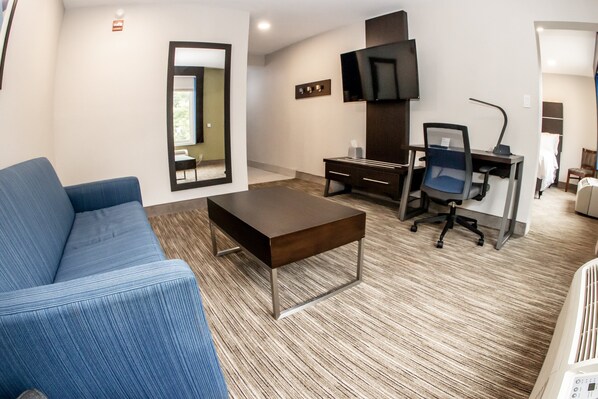 A modern hotel room with a blue sofa, a wooden coffee table, a desk with a chair, a flat-screen TV, and a large mirror. - Holiday Inn Express Allentown North by IHG (Allentown)