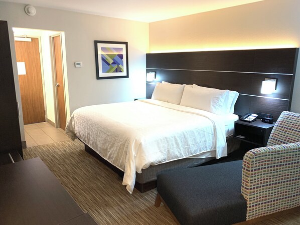 A hotel room with a bed, bedside table, chair, and a framed picture on the wall.