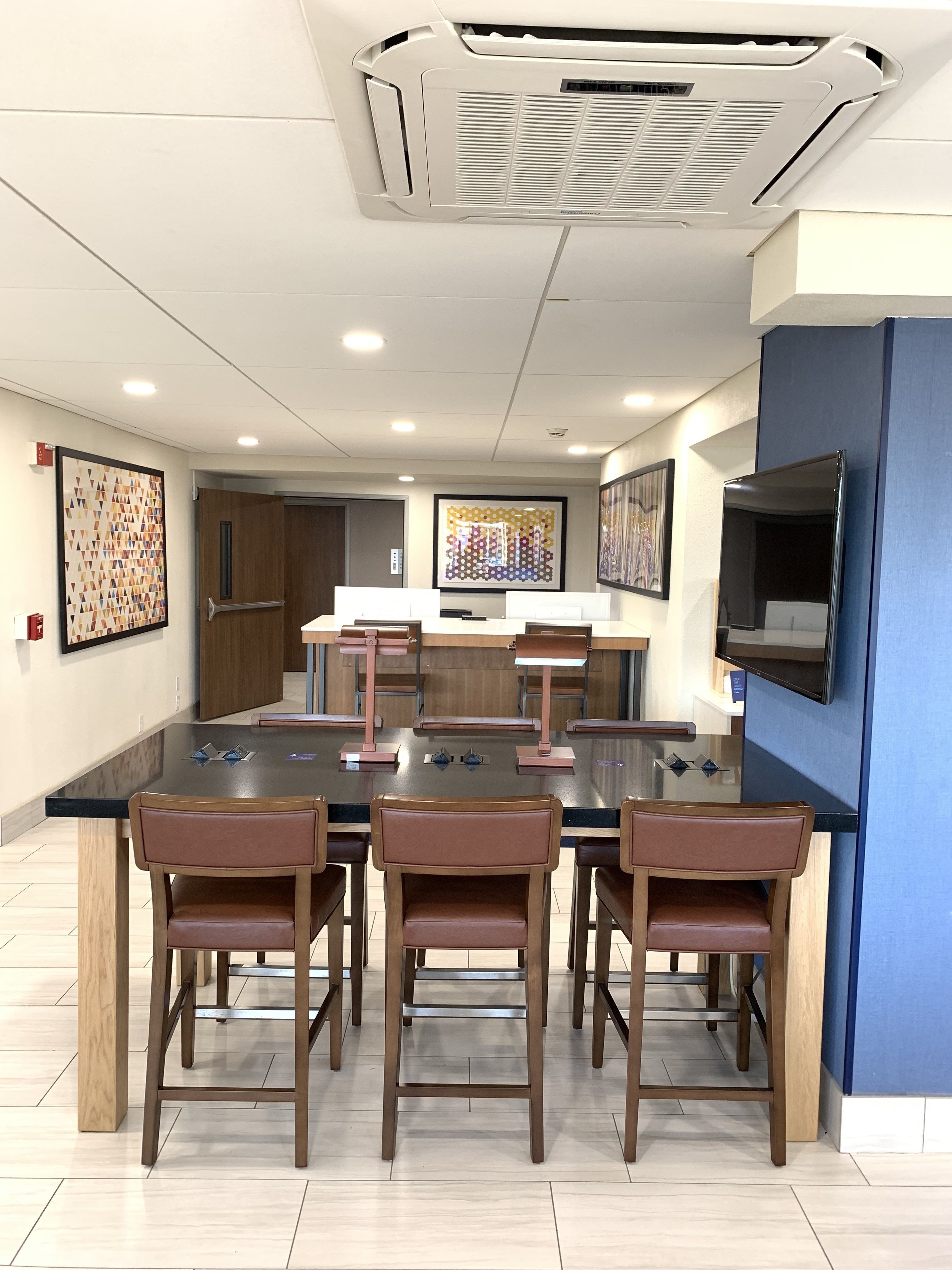 A modern dining area with a black table and six brown chairs. There is a ceiling air vent and a mounted flat-screen TV on the wall.